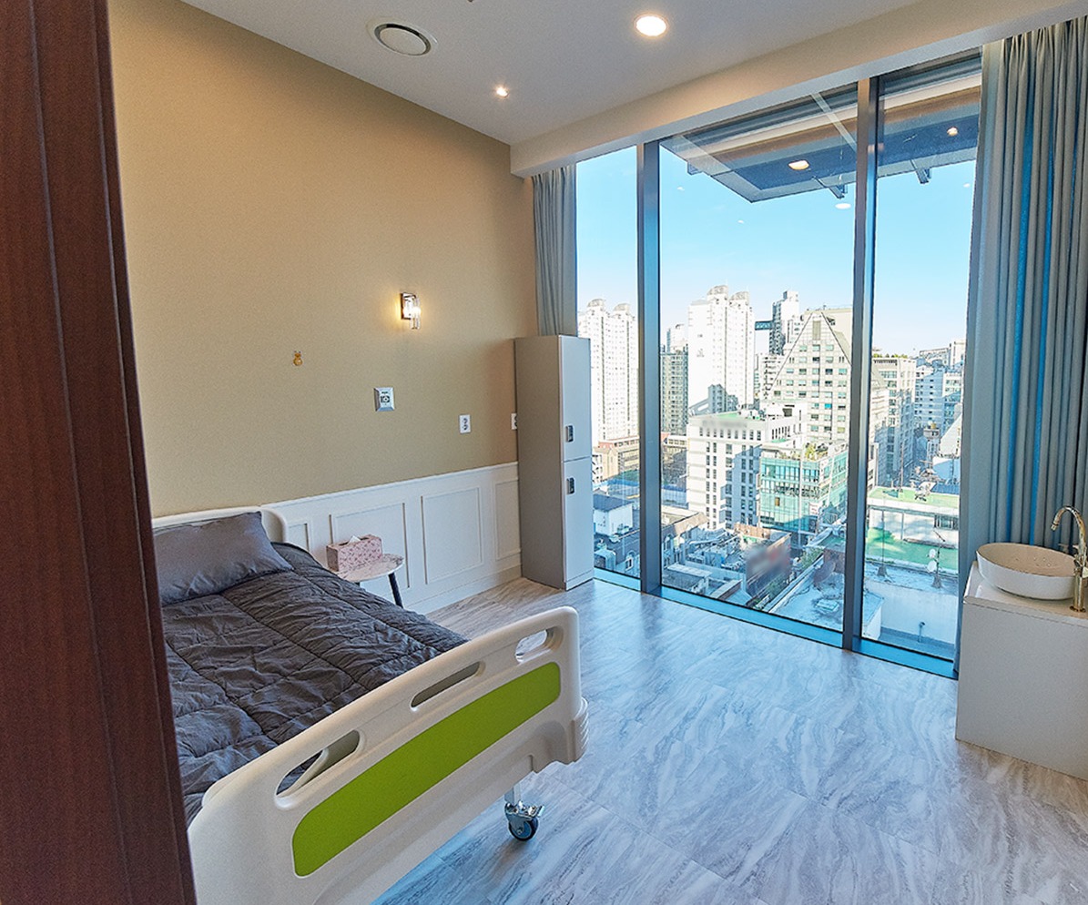 Private recovery room with a single bed, a small washbasin and floor-to-ceiling windows overlooking city buildings